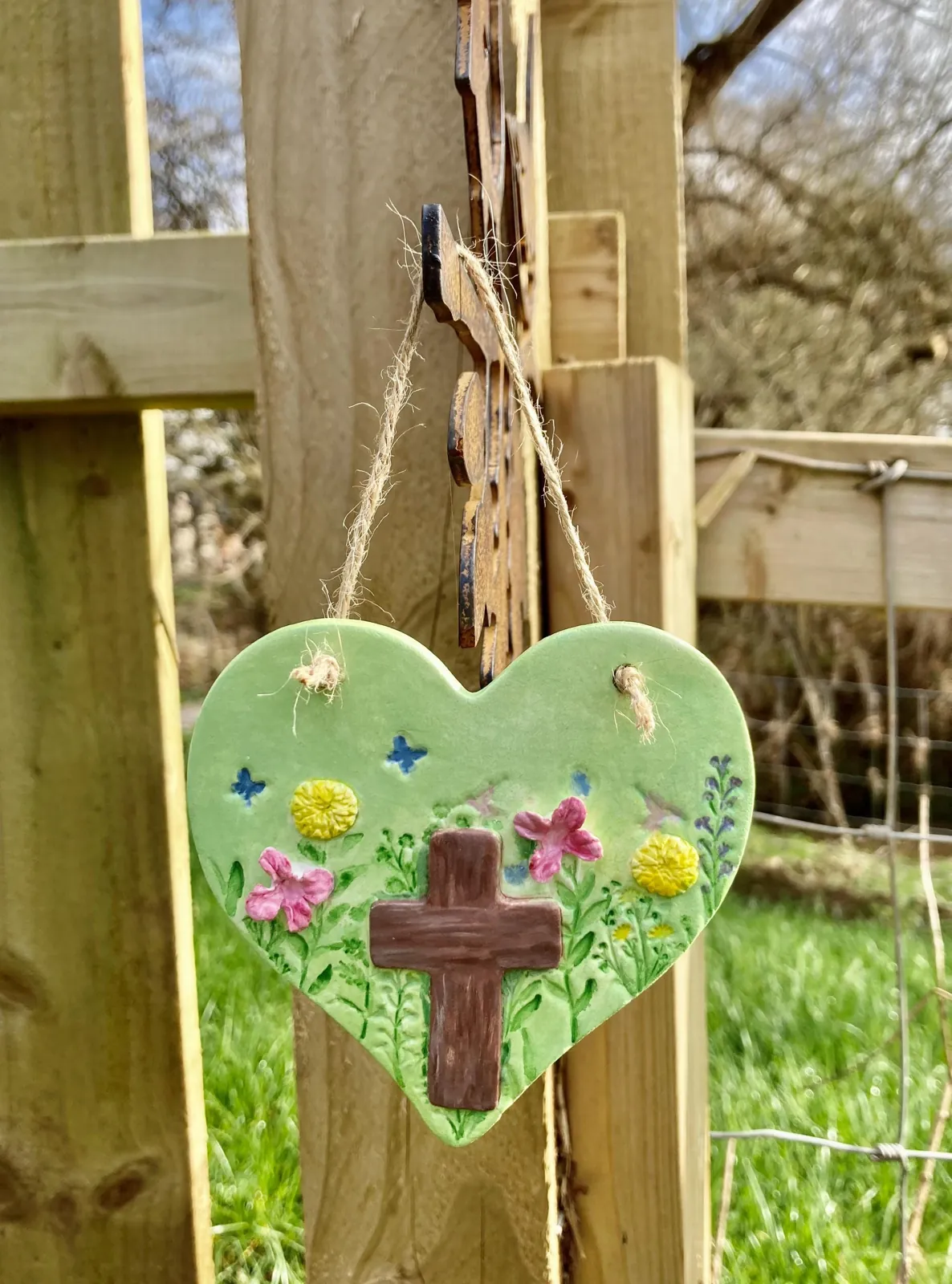 A green air-dry clay heart with a brown cross, butterflies and pink and yellow flowers, hanging by twine from a wooden fence post
